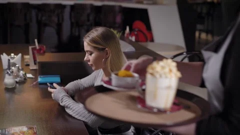 Blonde girl is sitting in cafe, using phone, waitress is bringing coffee and Stock Footage 81464245