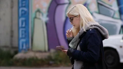 Blonde in glasses smokes an electronic cigarette and looks at  smartphone screen Stock Footage 130984138
