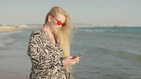 Blonde with glasses texting on smartphone while standing on the beach Видео 197456000