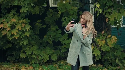 Blonde in gray coat doing selfies on smartphone in autumnal park Stock Footage 80694293