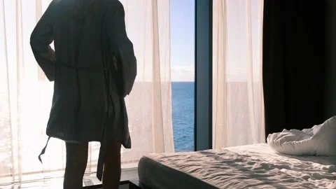 Blonde in a gray silk bathrobe, opens curtains on the window, view of sea and Stockbeeldmateriaal 165408026