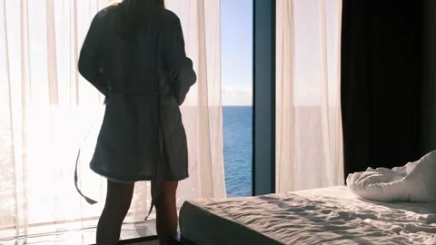 Blonde in a gray silk bathrobe, opens curtains on the window, view of sea and Stock Footage 165518453