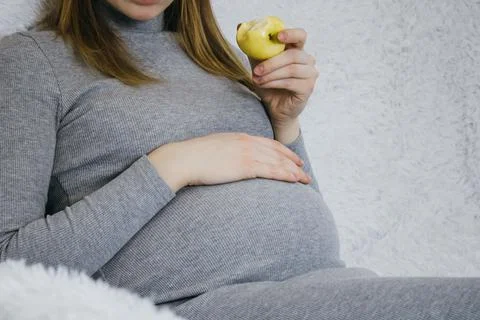 A blonde pregnant girl in a gray dress is sitting on a white sofa and eating  Stock Photos