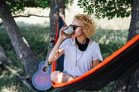 Blonde sitting in a hammock, in a shadow, between two big trees. Drinking tea Stock Photos