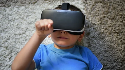 Blonde small boy lying on the carpet using VR headset and gesticulating Stock Footage 250364134