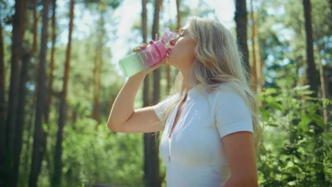 Blonde woman drinks from bottle while walking through tall pine trees Stock Footage 346663060