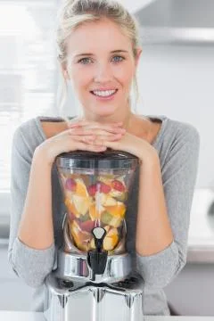 Blonde woman leaning on her juicer full of fruit and smiling at camera Stock Photos