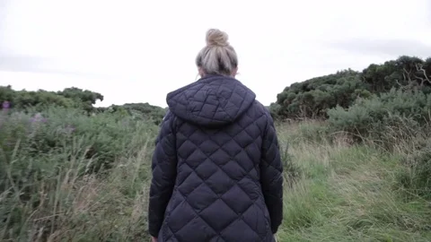 Blonde woman walks on path through bushes close to the ocean Stock Footage