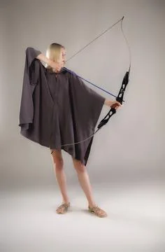 Blondie in short cloak and sandals aiming with a modern bow on gray backgrou Фото