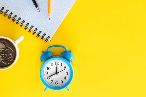Blue alarm clock, notepad and cup of coffee on a yellow background, flat lay. Stock Photos