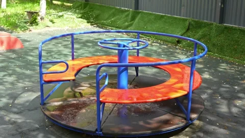 Blue and orange playground carousel spins slowly in dappled sunlight Stock Footage 335580545