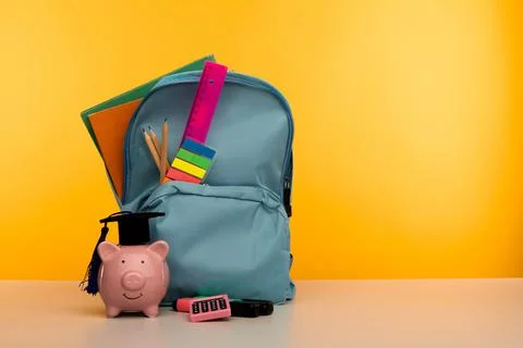 Blue backpack with school tools and piggy bank in hat on yellow background Stock Photos