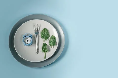 Blue clock, fork and spinach leaves on gray plate, intermittent fasting diet Stock Photos
