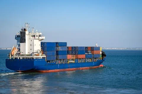 Blue container ship loaded with cargo containers sailing on sunny day near Stock Photos