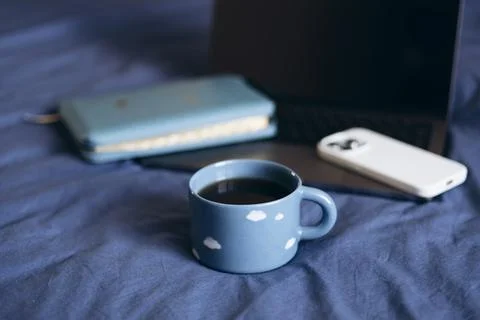 Blue cup with clouds on blue bed with blurred Bible, laptop and smartphone. Stock Photos