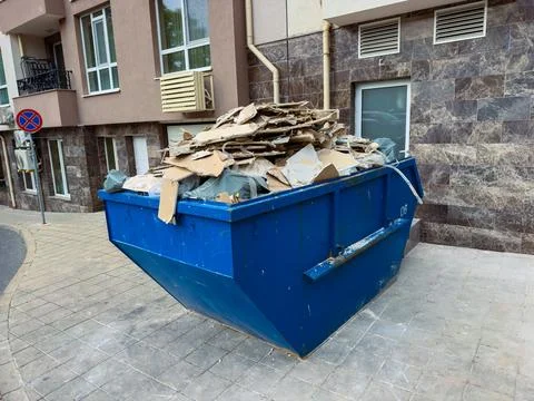 A blue dumpster sits on a paved area next to a building. Cardboard and constr Stock Photos