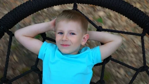 The blue-eyed five-year-old boy lies on a round swing with his hands behind his Stock Footage 89921566
