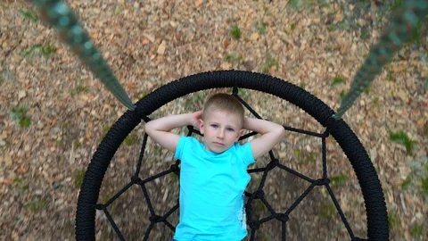 The blue-eyed five-year-old boy in blue t-shirt lies on a round swing with his Stock Footage 89921857