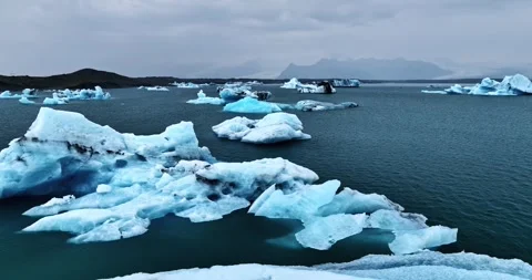 Blue icebergs float in a cold Icelandic lagoon, seen from the snowy shoreline 스톡 동영상 325163006
