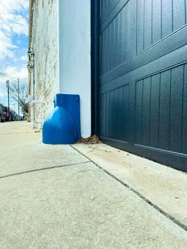 A blue object stands next to a dark garage door on a sidewalk. The scene show 库存照片