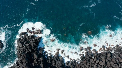Blue ocean waves break into white foam by black stones on a shore. Aerial of Stock Footage 117273808