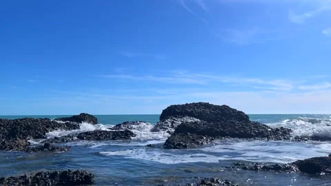 Blue ocean waves hitting dark rocks under sunny clear sky Stock Footage 310240291