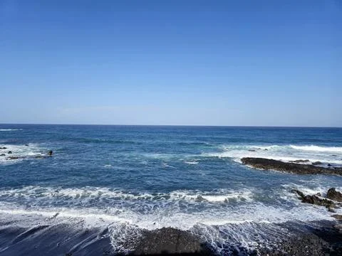 Blue ocean waves roll toward a rocky shoreline under a clear sky, creating .. Stock Photos