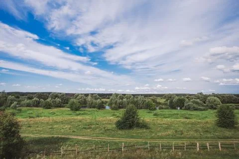 Blue sky with charming white clouds, green trees, meadow, river, rural road Stock Photos