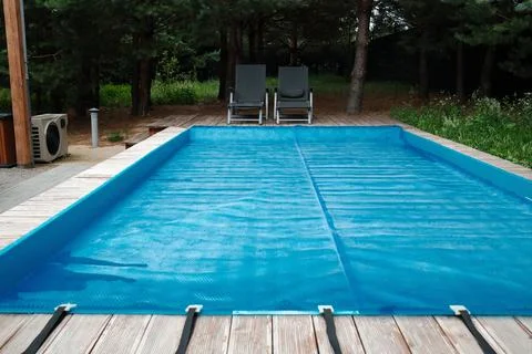 Blue solar pool cover on a backyard deck with chairs and pine trees 스톡 사진