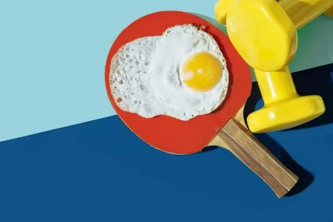 On the blue table are yellow dumbbells, a red ping pong racket, and eggs. Stock Photos