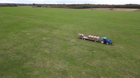 A blue tractor is pulling a fully loaded trailer of round hay bales across Stock Footage 287707484