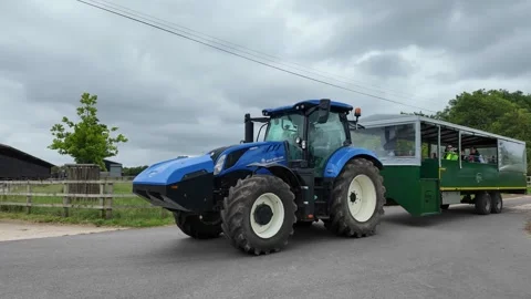 Blue tractor pulling a green passenger trailer on a farm road 库存影片 337872368