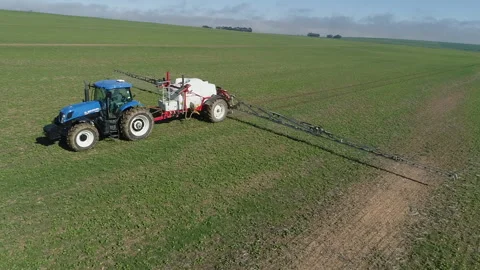 Blue tractor pulls agricultural sprayer over low green field, aerial Stock Footage 303884824