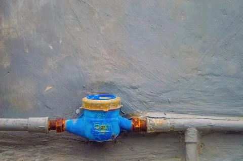 A blue water meter attached to a section of pipe against a textured gray wa.. Stock Photos