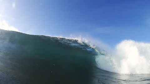 Blue wave forms hollow barrel beside rocky coast, underwater rear view at midday 库存影片 348702080