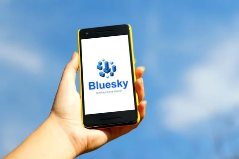 Bluesky app. Hand holding smartphone in yellow plastic case on blue sky nature Stock Photos