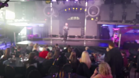 Blurred audience facing brightly lit stage in indoor event hall with pink purple Stock Footage 348157552
