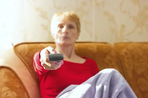 Blurred background. A TV remote control is in the foreground. An elderly woman Stock Photos