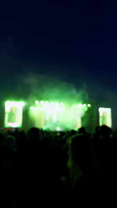 Blurred concert crowd facing bright outdoor stage lights at night with colorful Stock Footage 345763225