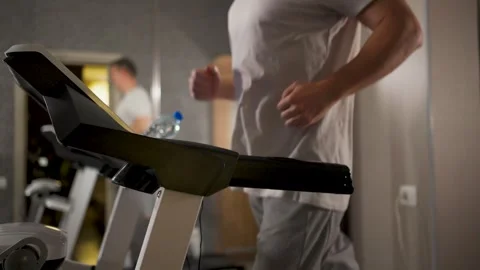Blurred shot of a man in a white t-shirt and grey shorts running on a tread.. 스톡 동영상 332054812