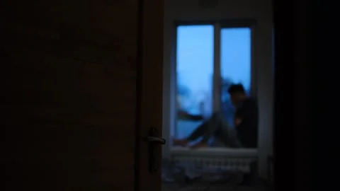 Blurred view of a man sitting on a windowsill, seen through an open door, convey Stock Footage 296425030