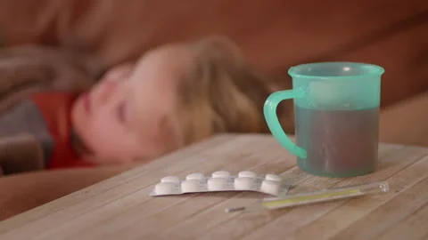 Blurred view of sick child lying on the sofa, hot drink, pills and thermometer Stock Footage 229608704