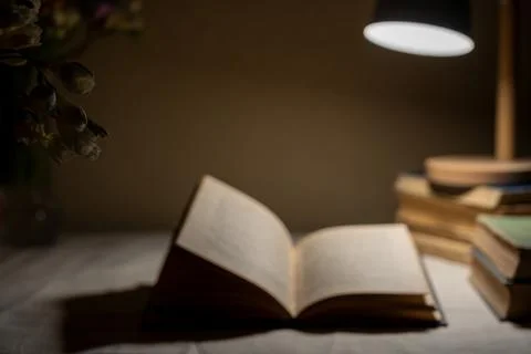 Blurry open book and stack of old books illuminated by lamp. Stock Photos