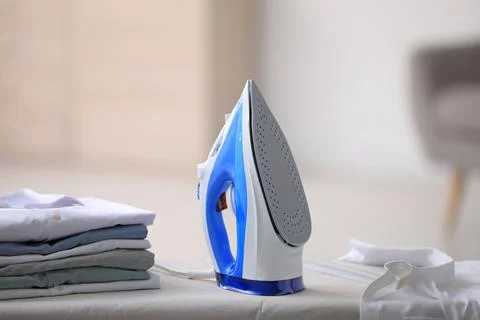 Board with modern iron and stack of clothes indoors. Laundry day Stock Photos