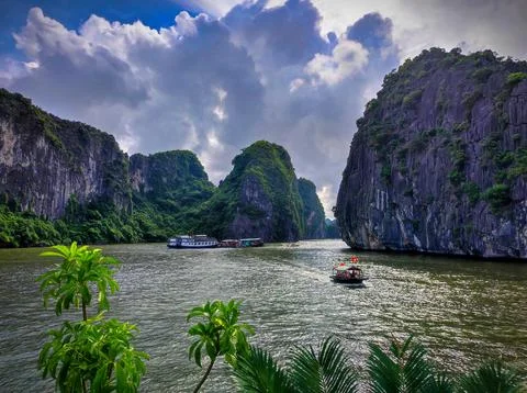 Boats navigate through green cliffs and water in a bay with clouds and sky .. Stock Photos