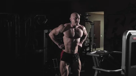 Bodybuilder posing and demonstrating chest muscles and arms in dark gym Stock Footage 99371635