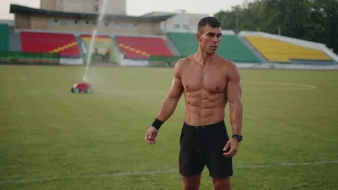A bodybuilder stands under drops of water from a sprinkler on a football field Vídeo Stock 162379477