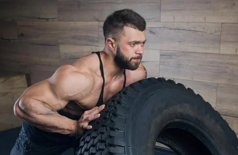 A bodybuilder with a tattoo and beard is pushing a tire in a gym. Stock Photos