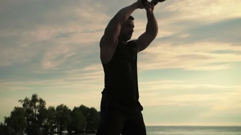 Bodybuilder is training outdoors, throwing kettlebells in front on sunrise.  Stock Footage 200118072