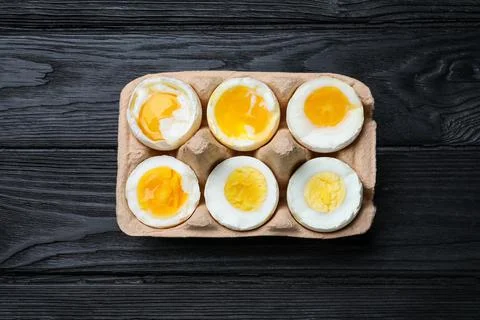 Boiled chicken eggs of different readiness stages in carton on black wooden t Stock Photos
